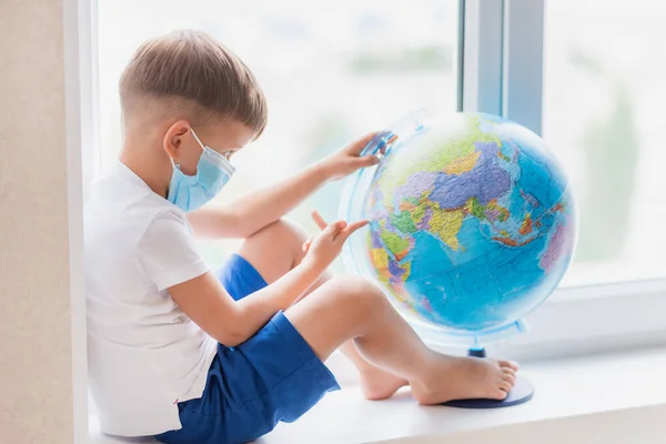 The masked kid sits at home in quarantine. The child is examining the globe while sitting on the windowsill. Prevention of coronavirus Covid-19 during a pandemic.