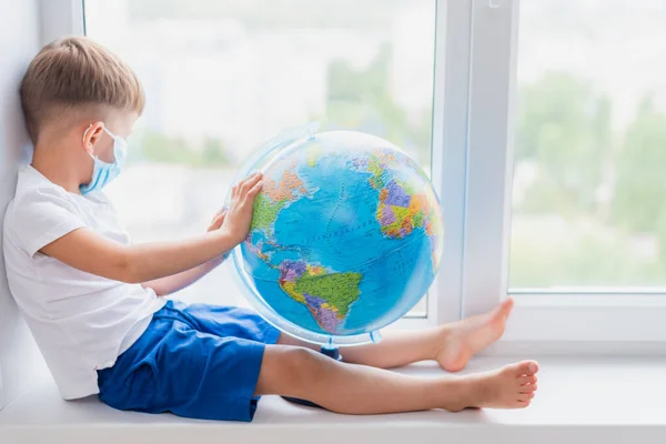 A child in a mask sits at home in quarantine. The child studies the globe while sitting on the windowsill. Prevention of coronavirus Covid-19 during a pandemic.