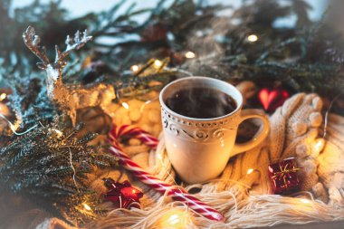 Christmas still life with a white cup and hot tea, with a white knitted scarf and New Year's decor against the background of spruce branches.