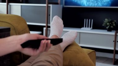 Kanepede uzaktan kumandayla televizyon programlarını değiştirmeyi kapat. Beyaz çoraplı bacaklar televizyonun önünde yatıyor. Ayaklarına yakın çekim. Modern odanın rahat atmosferi..