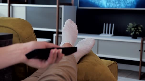 Primer plano de la conmutación de programas de televisión sentado en el sofá utilizando el control remoto. Las piernas en calcetines blancos yacen frente al televisor. Primer plano de sus pies. Ambiente acogedor de la habitación moderna. — Vídeos de Stock