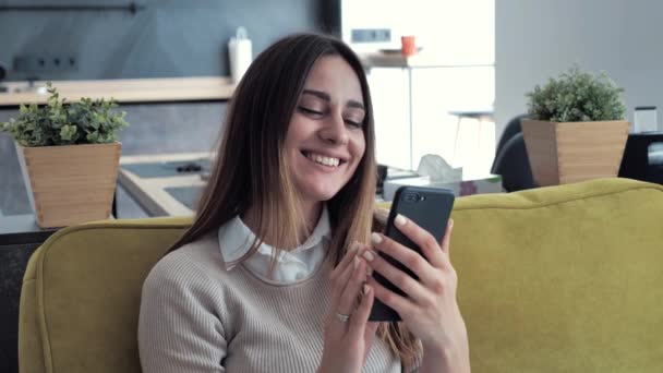 Расслабленная молодая женщина, улыбающаяся, используя смс-ки со смартфона, проверяя новости, просматривая социальные сети, сидя на диване. Тысячелетняя леди проводит время дома с клеточной технологией. 4k — стоковое видео