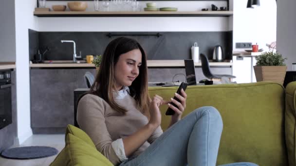 Belle jeune femme dans un pull beige assis dans le canapé en utilisant des messages texte de téléphone intelligent, vérifier les nouvelles, surfer sur les médias sociaux, assis sur le canapé. Millennial dame passer du temps à la maison avec la cellule — Video