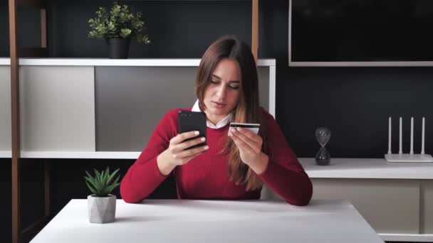 Sorridente giovane cliente donna in possesso di carta di credito e smartphone seduto in ufficio. Acquirente femminile felice utilizzando istantanei pagamenti mobili facili fare l'acquisto in negozio online. E-banking app service 4k — Video Stock