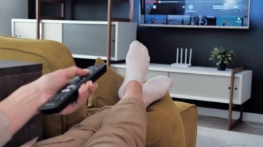 El Bas Tuşlara bas Gümüş Uzaktan Kumandalı TV Kontrolörü, koltukta dinlenen ayaklar 4k numaralı oturma odasında rahatça rahatlamak için televizyon seyrediyor.