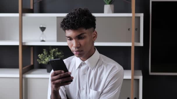 Joven con estilo Millennial Boy en camisa blanca usando su Smartphone para chatear, escribiendo mensajes mientras que de fondo interior moderno 4k — Vídeo de stock