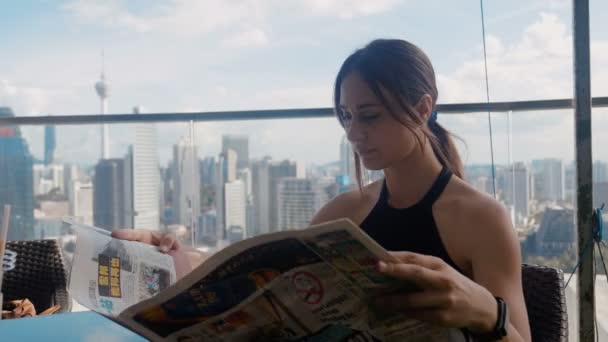 Ocupada mujer de negocios sentada en una cafetería bebiendo café con leche y leyendo un periódico el fondo de rascacielos 4k — Vídeos de Stock