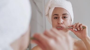 Kafasında havlu olan çekici bir kadın aynanın önünde dişlerini fırçalıyor.