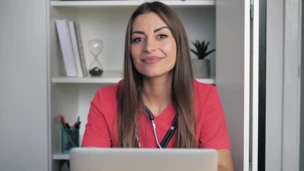 Giovane medico donna pensieroso indossa l'uniforme medica, si siede in clinica in ufficio con il computer portatile gira lo sguardo alla macchina fotografica, sorridente amichevole. Ritratto di infermiera 4k — Video Stock