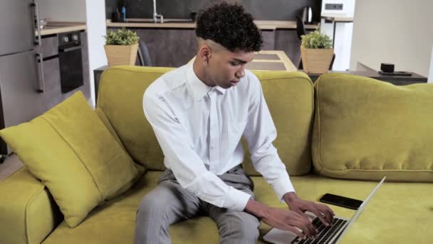 Joven hombre afro freelancer utilizando el dispositivo portátil en el sofá acogedor en la oficina en casa, retrato de un hombre que trabaja a distancia en el ordenador en el interoir moderno 4k — Vídeo de stock