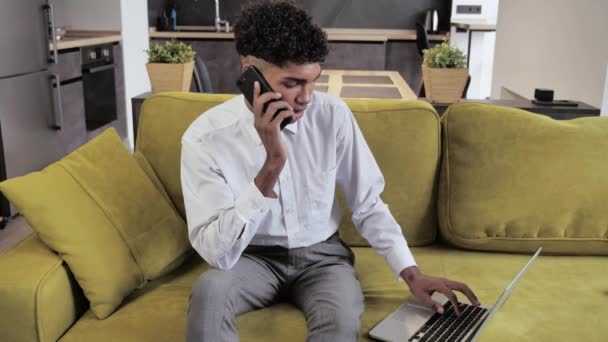 Κοντινό πλάνο νεαρός Αφρικανός Χρησιμοποιώντας φορητό υπολογιστή και κάνει τηλεφωνήματα στο γραφείο του σπιτιού. Του παίρνει περισσότερη δουλειά γίνεται στο ήμισυ του χρόνου 4k — Αρχείο Βίντεο