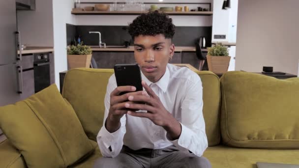 Forstyrret frustreret ung sort mand læser dårlige nyheder på smartphone. Trist fyr, der bruger telefonen derhjemme. Træt blandet race fyr sidder på sofaen føles smerte i øjnene 4k – Stock-video