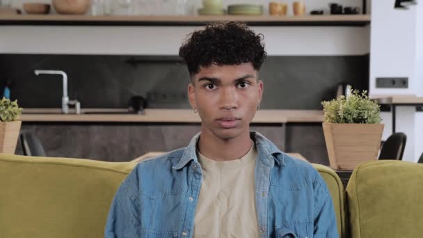 Vrolijke zwarte jongeman zit op een groene bank en glimlacht tegen een moderne interieur achtergrond. 4k — Stockvideo