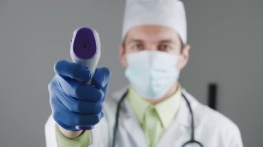 Sağlık çalışanı, doktor önlüğü giymiş, vücut ısısını ölçmek için kızıl ötesi termometreyle temas kurmayan birini tutuyor ve test ediyor. Sağlık ve tıbbi tema. Hastanede vücut ısısı kontrolü.