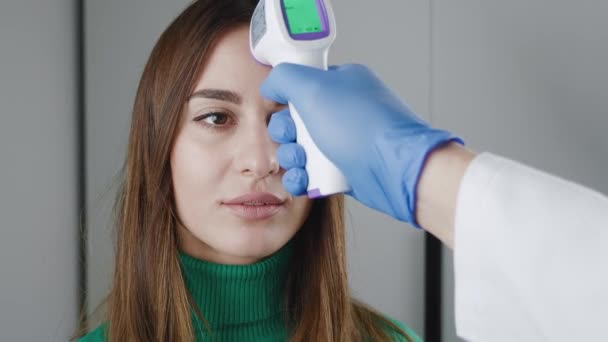 Medición de temperatura por termómetro electrónico infrarrojo para una mujer joven en suéter verde. Mujer con fiebre alta. Un control obligatorio en el trabajo con una pistola de termómetro es una frente sin contacto — Vídeos de Stock