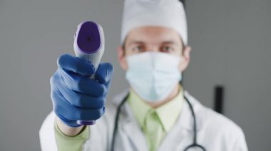 Sabahlık ve maske takmış bir erkek doktorun bulanık silueti. Temas olmayan termometreye odaklan.