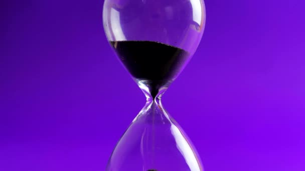 Tournage d'une horloge à sable mesurant le temps pendant que le sable tombe sur le fond violet - vieille minuterie classique. Extrême gros plan d'un sablier transparent avec sable noir coulant - concept de temps — Video