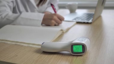 Arka planda çalışan kadın doktorla temassız kızılötesi tıbbi ekipmanların yakın görüntüsü. Masada dizüstü bilgisayar kullanarak notlar yazan beyaz önlüklü profesyonel bir doktor. Genel