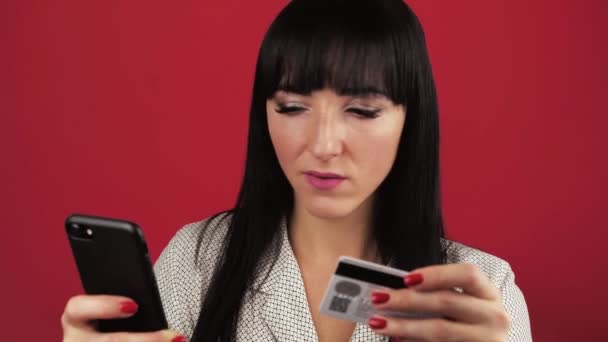 Mujer joven sosteniendo un teléfono inteligente en sus manos y haciendo compras en línea a través de una tarjeta de débito sobre fondo rojo. — Vídeos de Stock