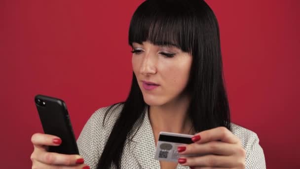 Σκούρα μαλλιά νεαρή γυναίκα 30 ετών κάνει μια αγορά μέσω του Διαδικτύου, ενώ κατέχει ένα smartphone και μια πιστωτική κάρτα πάνω από το κόκκινο φόντο — Αρχείο Βίντεο