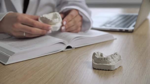 Orthodontiste médecin assis espace de travail indique un problème avec les dents sur le lingot de plâtre maxillaire lecture de l'anatomie du livre dans le fond flou. Concept de pratique générale — Video