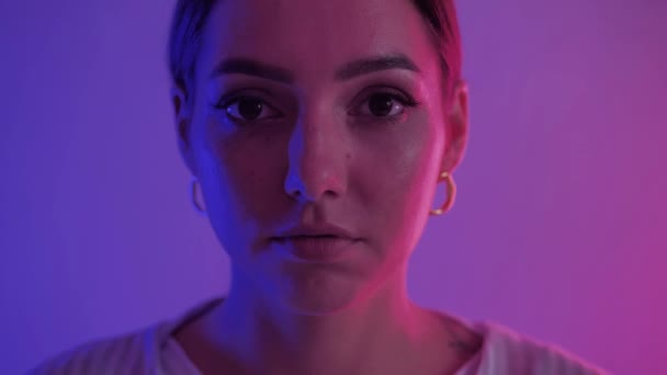 Neon-effect. Close-up portret van aantrekkelijke jonge brunette vrouw met oorbellen. Ernstig slim vrouwtje kijkt direct naar camera onder neon licht tegen wazig violette achtergrond — Stockvideo