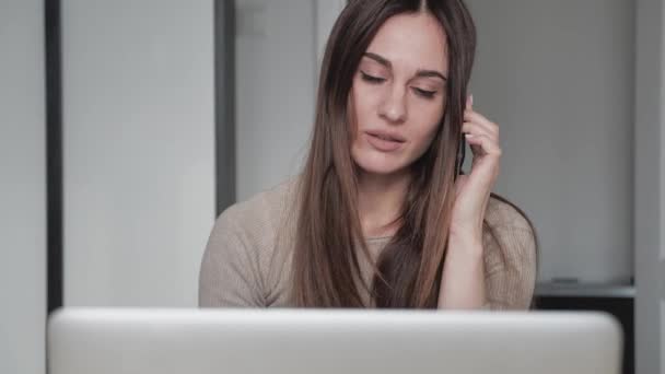 Komoly üzletasszony, aki laptoppal beszél mobilon, ül az asztalnál. Szakmai női menedzser tanácsadás ügyfél online projekt üzleti hívás a munkahelyen. — Stock videók