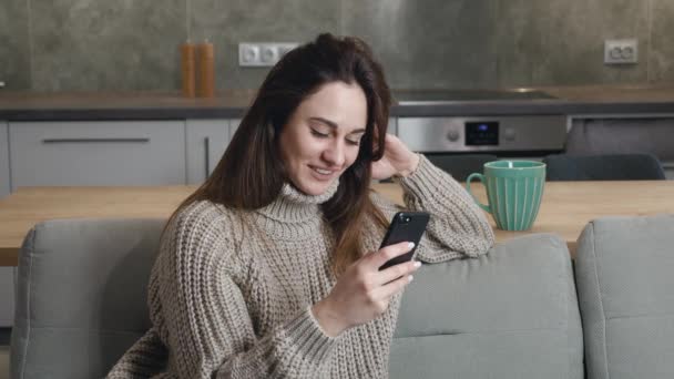 Mujer joven relajada en una sudadera de punto gris sonriendo usando mensajes de texto de teléfonos inteligentes, revisando noticias, navegando por las redes sociales, sentada en un sofá acogedor. Milenial dama pasar tiempo en casa con la celda — Vídeos de Stock