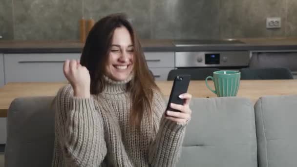 Πορτρέτο της ευτυχούς χαριτωμένο γυναίκα σε ένα μάλλινο πουλόβερ απολαύσετε την επιτυχία στο κινητό τηλέφωνο στο σπίτι kithen. Closeup χαρούμενη χιλιετή θηλυκό ανάγνωση καλά νέα ή να κερδίσει στο παιχνίδι στο τηλέφωνο. Έκπληκτη κυρία γιορτάζει — Αρχείο Βίντεο