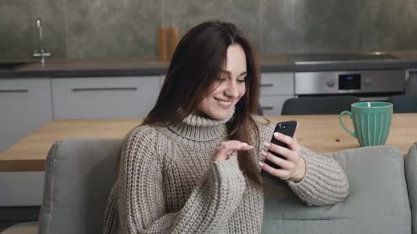 Giovane donna caucasica, 25 anni, si siede a casa su un divano in un maglione caldo e sfoglia rapidamente le pagine Internet sullo smartphone su un appartamento moderno. Focus selettivo, profondità di campo poco profonda — Video Stock