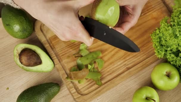 As mulheres descascam kiwi. Quivi de abacates fatiado e maçãs na mesa. cortes e cascas kiwi fresco suculento para uma salada de frutas. Dieta, poucas calorias, vegetarianismo, vitaminas naturais. — Vídeo de Stock