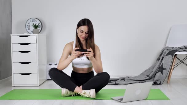 Молодая спортсменка сидит на коврике для йоги после онлайн тренировки. Женский холдинг, серфинг в Интернете, смотреть новости, играть в игры, общаться с друзьями глядя на ее телефон — стоковое видео