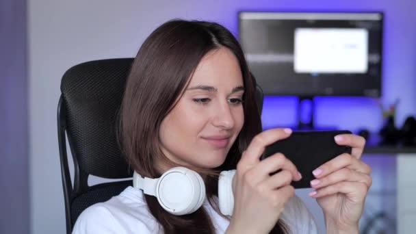 Femme gamer mignon assis sur une chaise de jeu, souriant et jouer jeu en ligne mobile sur un smartphone. Avoir du plaisir à jouer avec des amis en ligne. — Video