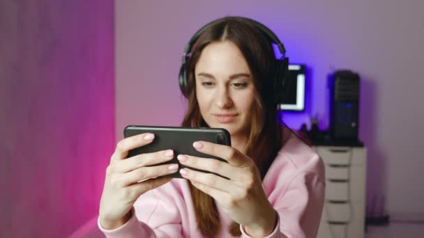 Χαριτωμένο gamer γυναίκα κάθεται σε μια καρέκλα τυχερών παιχνιδιών, χαμογελώντας και παίζοντας κινητό online παιχνίδι σε ένα smartphone. Διασκεδάζοντας παίζοντας με τους φίλους σε απευθείας σύνδεση. Έννοια νίκης — Αρχείο Βίντεο