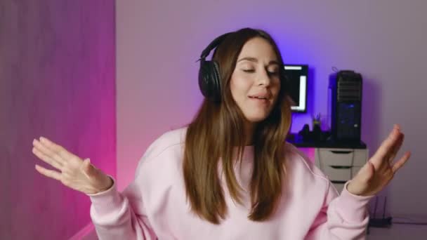 Молода жінка в рожевому светрі в навушниках танцює і співає в сучасній кімнаті з неоновим фоном — стокове відео