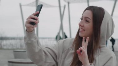 Bej kapüşonlu neşeli genç bir kadının akıllı telefondan selfie çekişini ve dışarıda el kol hareketlerini yakından izleyin.