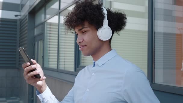 Mann mit Kopfhörer blickt auf Smartphone in der Nähe eines Gebäudes in der Stadt — Stockvideo