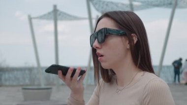 Güneş gözlüklü, neşeli, genç bir kadının akıllı telefondan gülümseyerek sesli mesajını kaydettiği yakın plan. Çekici bir kadının portresi.