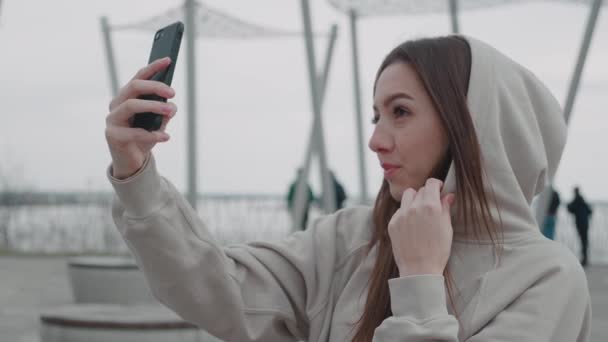 Attraktiv brunett hona i huva gör en selfie utomhus. Livsstilskoncept — Stockvideo