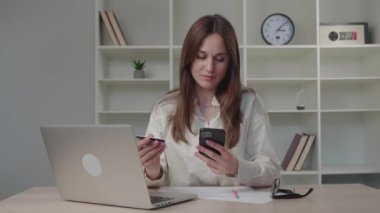 Elinde kredi kartı ve akıllı telefon olan gülümseyen genç bayan müşteri ev ofisinde oturuyor. İnternetten alışveriş yapan mutlu bayan alışverişçi kolay taşınabilir ödemeler yapıyor. E-bankacılık uygulama servisi