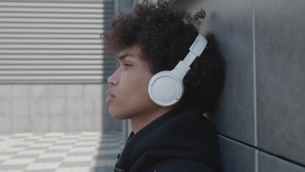 Kulaklıklı mutsuz bir Afro-Amerikan turist dışarıda müzik dinleyip sokakta akıllı telefon tutarak eğleniyor. Gençlik kültürü ve cihazları kavramı. — Stok video