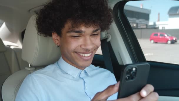 Joven chico de pelo rizado mixto leyendo noticias en Internet, jugando juegos en su teléfono inteligente, sonriendo lindo mientras está sentado dentro del coche — Vídeo de stock