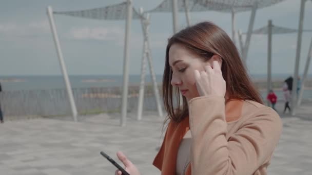 Atraktivní mladá žena poslech hudby ve sluchátkách pomocí smartphone ve městě chůze rozhlédnout úsměv portrét close up slow motion — Stock video