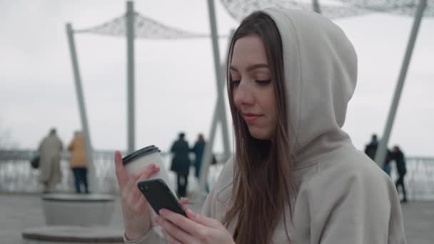 Mladá dívka v mikině pije kávu a listy přes zprávy krmení na smartphone venku — Stock video
