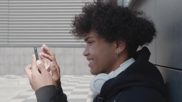 L'adolescente afro-americano aumenta le dimensioni dello schermo, sfogliando il feed di notizie, facendo un acquisto online, giocando a giochi per smartphone e ascoltando musica vicino alla costruzione — Video Stock