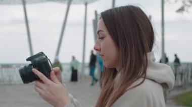 Elinde fotoğraf makinesi olan ve bir objeye odaklanan genç bir kadın gülümsüyor ve parkta fotoğraf çekiyor.