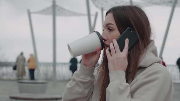 Caucásico alegre joven elegante mujer hablando por teléfono celular y bebiendo bebida caliente por la mañana en la calle. Hermosa mujer feliz hablando por teléfono móvil y tomando café. Afuera.. — Vídeo de stock