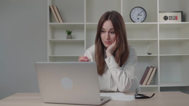 Young serious girl working in a modern office on a laptop, typing, writing a note in a notebook — Stock Video
