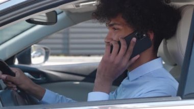 Genç siyah bir erkek, kulağına cep telefonu dayamış arabada oturuyor..