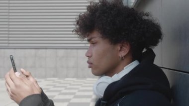 Siyah kapüşonlu mutlu Afro adam kulaklıklı müzik dinlerken cep telefonuyla internetten mesajlaşıyor.